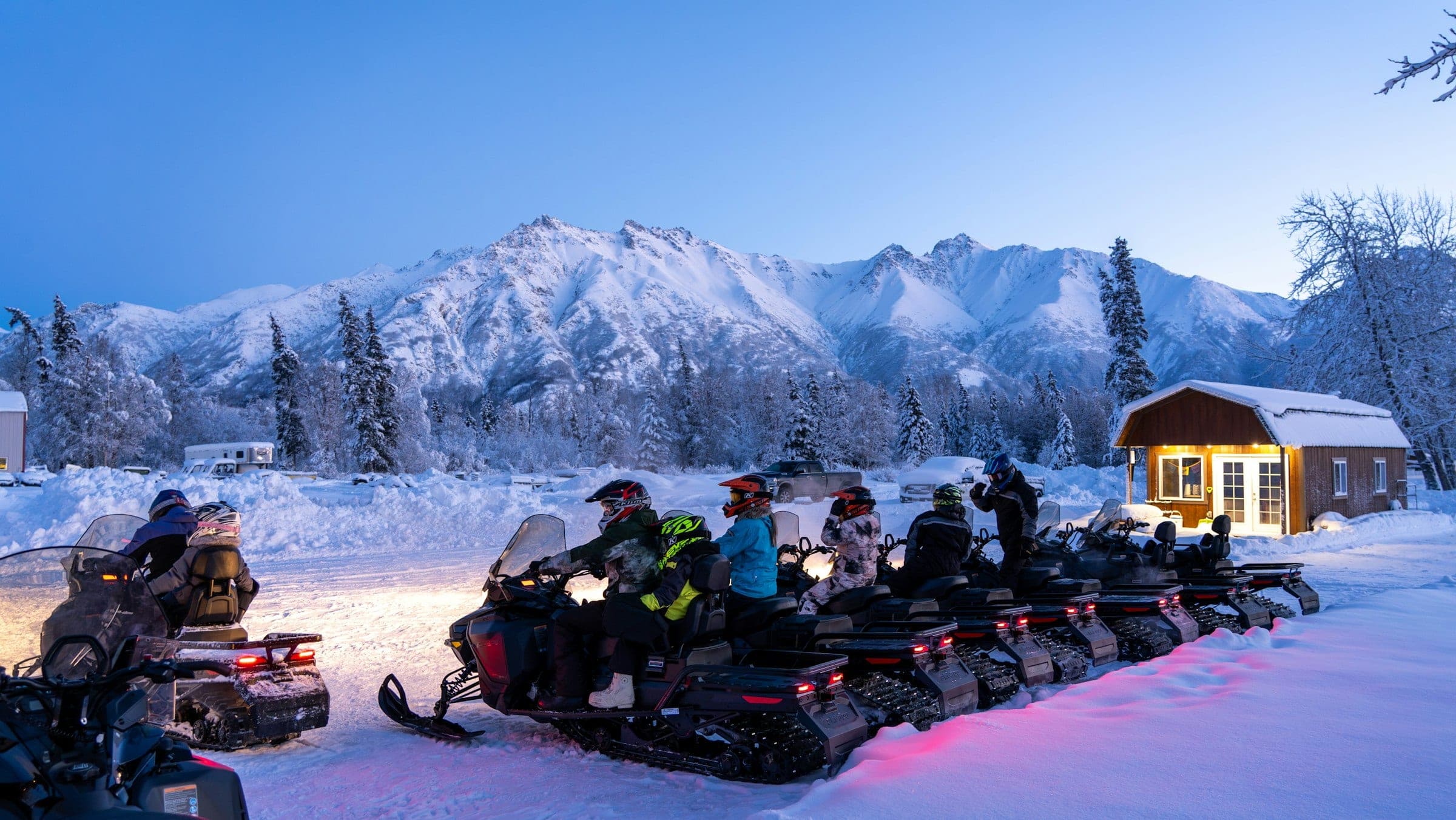 A group setting out on snowmobiles at blue hour, snowy peaks behind them